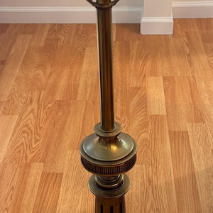May include: A tall brass table lamp base featuring a cylindrical column with vertical grooves, a tiered circular base, and a metal harp assembly at the top. The lamp includes a central socket with a turn-knob switch and a black electrical cord.