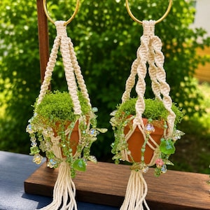 Pode incluir: Dois vasos de terracota em miniatura com musgo verde brilhante, suspensos em suportes de macramé em tom off-white com detalhes de corda torcida. Cada suporte apresenta contas de vidro verde, detalhes em forma de folha e pequenas flores.