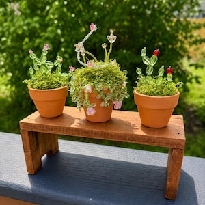 Pode incluir: Três vasos de terracota em miniatura contendo musgo verde e esculturas de plantas feitas de contas. O vaso da esquerda apresenta caules de contas verdes com detalhes em rosa. O vaso central tem vinhas e flores de contas pendentes. O vaso da direita.