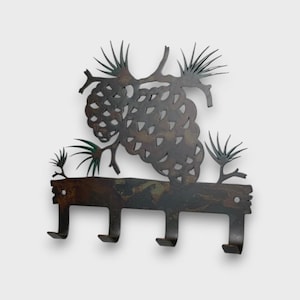 May include: A dark brown metal wall hook rack featuring a decorative cutout design of a pinecone with pine needles. The horizontal base includes four downward-curving hooks for hanging items, with two circular mounting holes on the top corners.