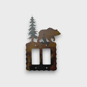 May include: A double light switch plate cover made of metal with a rustic, mottled brown and black finish. The top edge features a silhouette cutout of a standing bear and a tall pine tree. The plate has two rectangular openings for switches.