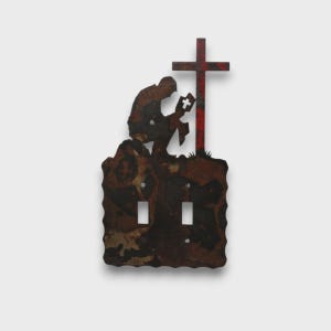 May include: A double light switch cover plate made of dark, weathered metal with a rusted finish. The plate features a silhouette of a person kneeling in prayer before a cross. The cross is accented with a red finish and a small cutout cross shape.