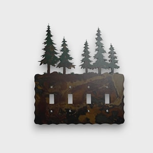 May include: A four-gang light switch cover plate featuring a cutout silhouette of five evergreen trees along the top edge. The rectangular base has a wavy bottom edge and a mottled, multi-toned brown and black finish resembling natural stone.
