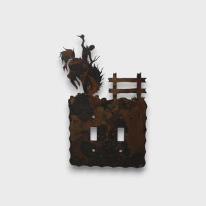 May include: A double light switch plate made of dark, rusted metal with a scalloped edge. The design features a silhouette of a cowboy riding a bucking horse next to a wooden fence post. The plate includes two rectangular cutouts for switches.