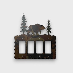 May include: A rectangular metal light switch cover plate with four vertical rectangular openings. The plate features a dark, mottled brown and black finish with a silhouette of a bear walking between two pine trees extending from the top edge.