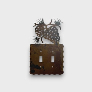 May include: A double light switch plate cover made of dark brown metal with a weathered, rustic finish. The rectangular base features a scalloped edge and two vertical toggle switch cutouts. A decorative cutout of pine cones and needles sits atop.