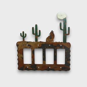 May include: A four-gang light switch cover plate featuring a rustic, mottled brown and black finish. The top edge displays a desert scene with three green saguaro cacti, a howling coyote silhouette, and a circular white moon against the wall.