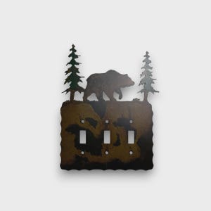 May include: A triple light switch cover plate made of dark brown metal with a textured finish. The top edge features a silhouette of a walking bear positioned between two evergreen trees. The plate has three rectangular cutouts for toggle switches.