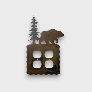 May include: A metal double outlet wall plate featuring a cutout silhouette of a bear walking past a pine tree. The plate has a mottled brown and dark metallic finish with four circular openings for electrical sockets and two small screw holes.