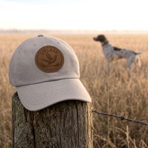 Puede incluir: Una gorra de béisbol de color beige claro con un parche circular de cuero marrón en la parte delantera. El parche muestra un gráfico en relieve de un ave en vuelo sobre el texto UPLAND. La gorra tiene una visera curva y ojales cosidos.