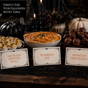 May include: Three white rectangular food label cards with black ornate borders and orange text reading Devilish Deviled Eggs, Pumpkin Dip, and Monster Meatballs. The cards are displayed in front of a bowl of orange dip, deviled eggs, and meatballs.