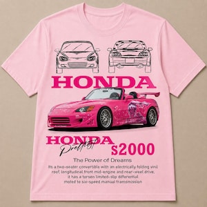May include: A light pink short-sleeve t-shirt featuring a graphic of a pink Honda S2000 convertible. The design includes line drawings of the car, the text HONDA S2000, The Power of Dreams, and a descriptive paragraph about the vehicle's specifications.