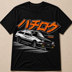 May include: A black short-sleeve t-shirt featuring a graphic of a white and black Toyota Sprinter Trueno AE86 car. Above the car is the orange Japanese text "ハチロク". To the right of the car, the text "SPRINTER TRUENO AE86" is printed in white and orange.