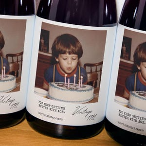 May include: Three dark glass wine bottles with white labels featuring a vintage photograph of a child blowing out candles on a birthday cake. The text reads: YOU KEEP GETTING BETTER WITH AGE. HAPPY BIRTHDAY JAMES! Vintage 1972.