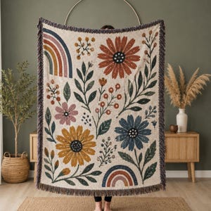 May include: A woven throw blanket featuring a cream-colored background with a repeating pattern of large rust, yellow, and blue flowers, green leaves, and small rainbows. The blanket has a textured knit construction and finished fringe edges.