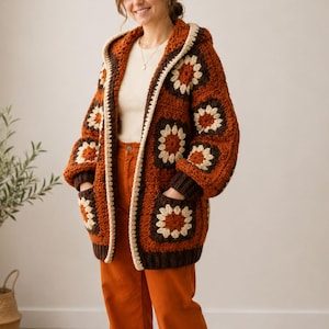 May include: A chunky crochet hooded cardigan featuring a granny square pattern in shades of burnt orange, dark brown, and cream. The garment has long sleeves, a front opening, and two large patch pockets, all finished with a cream-colored trim.
