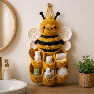 Puede incluir: Un organizador de pared de ganchillo con forma de abeja amarilla y negra, con una pequeña corona, alas blancas y seis bolsillos. Los bolsillos contienen artículos de aseo como botellas, un peine de madera y una esponja de baño blanca.