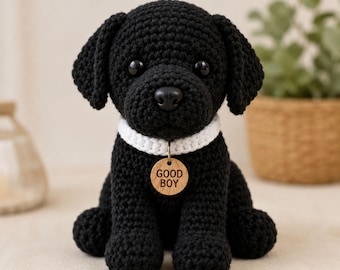May include: A black crocheted dog toy with floppy ears, black plastic eyes, and a black nose. The dog wears a white crocheted collar with a round wooden tag hanging from a metal ring. The tag is engraved with the text GOOD BOY in capital letters.