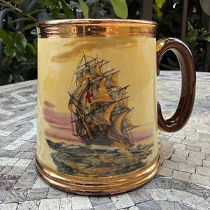 May include: A ceramic mug with a cream-colored body featuring a detailed illustration of a sailing ship with a red cross on its sails. The mug has a metallic gold rim and base, and a dark brown handle. The ship is depicted on choppy water.
