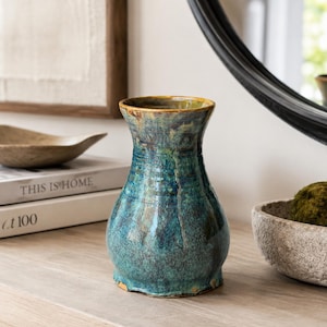 May include: A ceramic vase with a flared rim and a bulbous base, featuring a mottled glaze in shades of deep blue, teal, and brown. The surface has a glossy, textured finish with visible vertical drip patterns and subtle variations in color throughout.