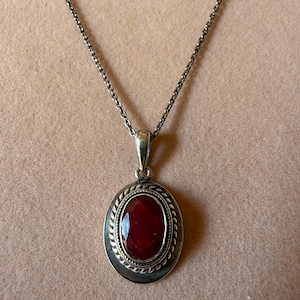 May include: A silver necklace featuring an oval pendant with a deep red, faceted gemstone at its centre. The pendant is framed by a twisted rope-style border and a smooth outer edge, suspended from a delicate silver chain with small links.