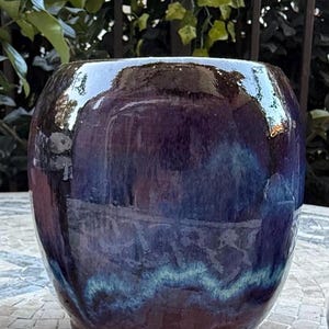May include: A rounded ceramic planter featuring a glossy, multi-toned glaze that transitions from deep purple and dark navy blue at the top to a lighter, vibrant teal-blue band near the base, with a smooth, reflective surface and a wide opening.