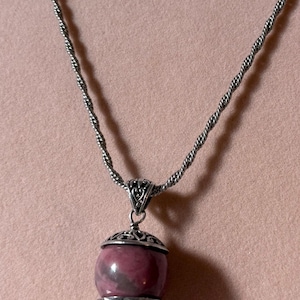 May include: A silver necklace featuring a twisted chain and a pendant with a round, polished, mauve-colored stone. The stone is held between two ornate, filigree-style silver caps, with a small silver bead at the bottom of the pendant structure.