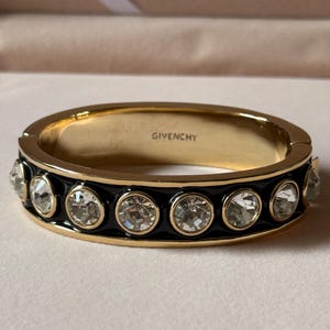 Może przedstawiać: Złota bransoletka typu bangle z zawiasem, ozdobiona czarną emalią oraz rzędem przezroczystych, okrągłych kryształów o szlifie fasetkowym w złotych oprawach. Wewnątrz bransoletki wygrawerowano nazwę marki GIVENCHY wielkimi literami.