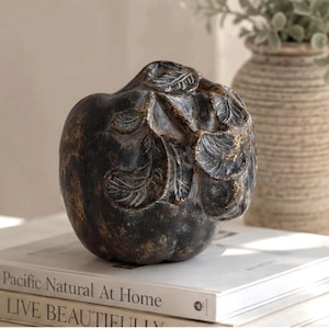 May include: A dark brown, apple-shaped decorative object with a textured, weathered finish and gold-toned highlights. The surface features sculpted, raised leaf details on the top section, creating a dimensional appearance with a matte, aged look.