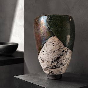 May include: A tall ceramic vase featuring a multi-textured finish with a glossy dark green and metallic bronze upper section, transitioning into a matte white lower base adorned with intricate black crackle patterns and a dark circular foot.