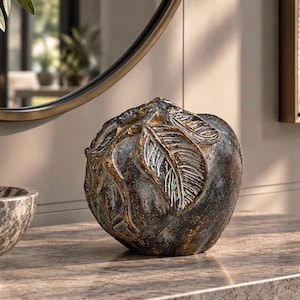 May include: A decorative apple-shaped sculpture featuring a dark, weathered finish with metallic gold highlights on the textured leaves and stem. The surface displays a distressed, multi-tonal appearance with deep charcoal and bronze undertones.
