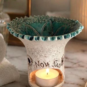 May include: A ceramic oil burner with a speckled off-white exterior and a vibrant teal interior. The flared top features a scalloped edge. The front displays the text Novi Sad above a circular opening containing a small, lit white tea light candle.