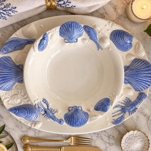 May include: A white ceramic dinner plate and matching bowl set featuring raised, three-dimensional blue-painted marine life motifs, including various seashells, a lobster, and a fish, arranged around the rims of both pieces.