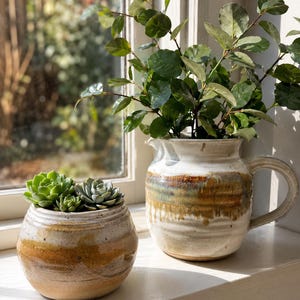 May include: A round ceramic planter containing green succulents and a ceramic pitcher with a handle holding a leafy green plant. Both vessels feature a white upper section and a textured, drip-style glaze in shades of brown and tan on the lower half.