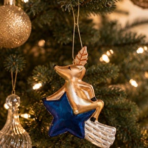 May include: A gold-coloured deer ornament sitting on a dark blue star with a white, grid-patterned tail. The deer has black accents on its hooves and ears, and a gold string loop at the top for hanging. The ornament is suspended from a tree branch.