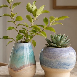May include: Two ceramic vases with a reactive glaze finish. The left vase is tapered with blue, teal, and beige tones, holding a green leafy plant. The right vase is rounded with purple, blue, and beige tones, containing a green succulent plant.