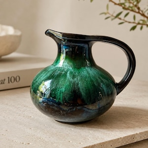 May include: A small ceramic pitcher with a rounded body and a curved handle. The glaze features a deep, glossy finish that transitions from a dark black rim down into a vibrant, mottled emerald green and blue center, ending in a dark base.