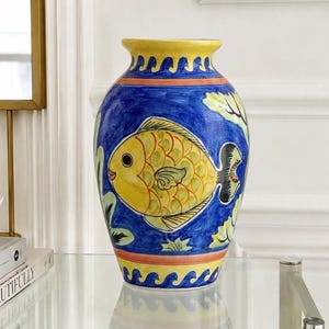 May include: A ceramic vase featuring a hand-painted yellow fish on a vibrant blue background. The vase has a yellow rim and base, both decorated with a blue wave pattern. The body of the vase displays green leaf accents surrounding the fish.