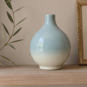May include: A round ceramic vase with a narrow neck, featuring a glossy finish that transitions from a pale blue at the top to an off-white base. The surface displays a fine, intricate crackle glaze pattern throughout the entire body of the vase.