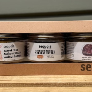 May include: A brown cardboard gift box with a rectangular cutout window containing three glass jars of Sequoia Nut Butter. The labels feature the text Carrot Cake Cashew Pecan Walnut Butter, Snickerdoodle Cashew Butter, and Pecan Pie Mixed Nut Butter.