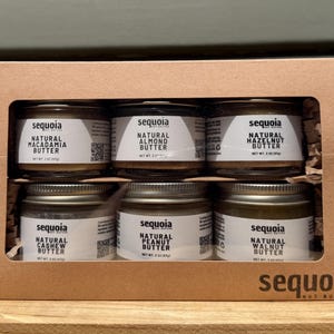 May include: A brown cardboard gift box containing six small glass jars of Sequoia nut butter. The jars are labeled Natural Macadamia, Almond, Hazelnut, Cashew, Peanut, and Walnut butter, each with a net weight of 2 ounces printed on the white labels.