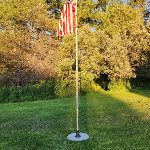 May include: A United States flag with red, white, and blue colours attached to a tall, thin, light-coloured metal pole. The pole is secured at the base by a circular, flat, light-grey stand resting on a green lawn with trees in the background.