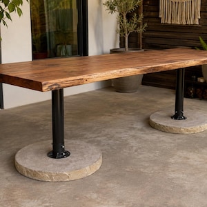 May include: A rectangular dining table featuring a thick, natural-edge brown wood slab top. The table is supported by two black metal cylindrical pedestals, each anchored to a heavy, circular, light-colored stone base resting on the ground.