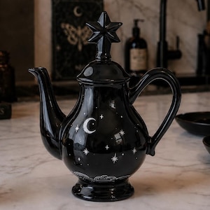 Op de afbeelding: Een glanzend zwarte keramische theepot met een wit patroon van een maansikkel en sterren op de ronde buik. Het deksel is bekroond met een stervormige knop. De theepot heeft een gebogen handvat en een tuit, rustend op een voetstuk.