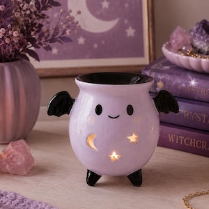 May include: A light purple ceramic wax warmer shaped like a round cauldron with black bat wings and three black feet. The front features a smiling face and cutouts in the shapes of a crescent moon and stars, with a warm light glowing from within.