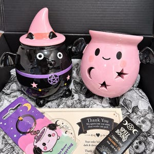 Könnte beinhalten: Ein schwarzer Keramikkessel mit einem rosa Hexenhut-Deckel, ein rosa Keramikkessel mit Stern- und Mond-Ausschnitten, ein Pastel Bat Brew Emaille-Schlüsselanhänger auf einer Karte und eine kleine schwarze Schachtel Spell Bound Frankincense Fragrance Oil 10ml.