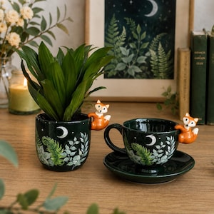 May include: A dark green ceramic planter and a matching teacup with a saucer, both featuring a white crescent moon, stars, and green fern leaf illustrations. Each piece has a small, three-dimensional orange and white fox figurine attached.