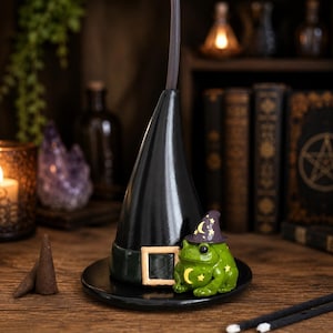 May include: A black incense burner shaped like a witch hat with a wide brim and a square buckle. A small green frog figurine wearing a purple hat with yellow stars and moons sits at the base of the hat. A thin incense stick protrudes from the top.