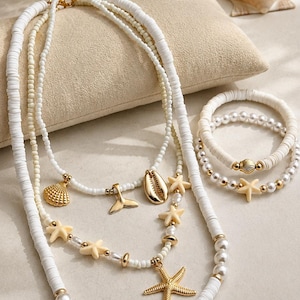 May include: A layered necklace set and two bracelets featuring white disc beads, small white pearls, and gold-toned metal accents. The jewellery includes charms shaped like a gold starfish, a gold seashell, a gold whale tail, and cream-coloured starfish.