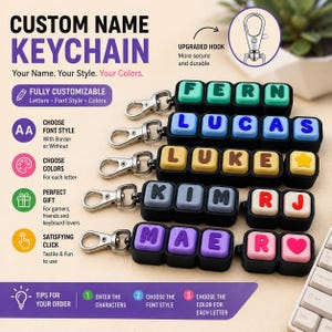 May include: Customisable name keychains featuring square, keyboard-style blocks with letters in various colours. Each keychain has a silver metal lobster clasp hook. The blocks are arranged in rows, displaying names like FERN, LUCAS, LUKE, KIM, RJ, MAE, and R.